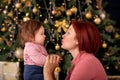 Mother holding hands and kissing infant baby daughter in front of decorated Christmas New Year fir tree. Family winter holidays Royalty Free Stock Photo