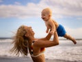 Mother holding and lifting her infant baby boy high in the air on beach. Positive emotions. Mum and son laughing and smiling. Blue Royalty Free Stock Photo