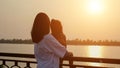 Mother holds little girl on railing on waterfront at sunset