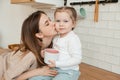 Mother kissing her daughter at home in kitchen, breakfast. Little girl looking at camera. Royalty Free Stock Photo