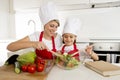 Mother and little daughter cooking together with hat apron preparing salad at home kitchen