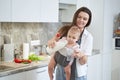 Happy cheerful mom with her cute baby in baby carrier preparing breakfast at home Royalty Free Stock Photo