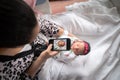 A mother in a pink animal print bathrobe sits on a hospital bed holding her newborn baby girl swaddled in a blanket while taking a