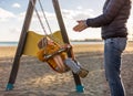 Mother pushing her infant baby boy child on a swing on sandy beach playground outdoors on nice sunny cold winter day in