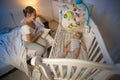 Young mother sitting on bed and looking on her baby son lying in cot Royalty Free Stock Photo