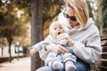 Mother sitting on bench in urban park, holding her infant baby boy child in her lap and feeding him baby food on nice Royalty Free Stock Photo