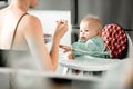 Mother spoon feeding her baby boy child in baby chair with fruit puree in kitchen at home. Baby solid food introduction Royalty Free Stock Photo