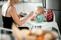 Mother spoon feeding her baby boy child in baby chair with fruit puree in kitchen at home. Baby solid food introduction Royalty Free Stock Photo