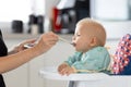 Mother spoon feeding her baby boy child in baby chair with fruit puree in kitchen at home. Baby solid food introduction Royalty Free Stock Photo