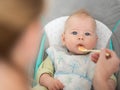 Mother spoon feeding her baby boy infant child in baby chair with fruit puree. Baby solid food introduction concept. Royalty Free Stock Photo