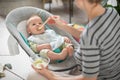 Mother spoon feeding her baby boy infant child in baby chair with fruit puree. Baby solid food introduction concept. Royalty Free Stock Photo