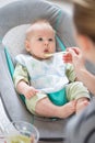 Mother spoon feeding her baby boy infant child in baby chair with fruit puree. Baby solid food introduction concept. Royalty Free Stock Photo