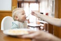 Mother spoon feeding her infant baby boy child sitting in high chair at the dining table in kitchen at home Royalty Free Stock Photo