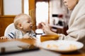 Mother spoon feeding her infant baby boy child sitting in high chair at the dining table in kitchen at home Royalty Free Stock Photo