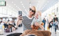 Mother taking selfie with mobile phone, while traveling with child, holding his infant baby boy at airport, waiting to