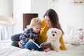 Mother, toddler child and puppy dog, lying on the bed, reading book together, family time