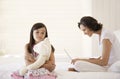 Mother Using Laptop With Daughter Holding Teddy Sitting On Bed