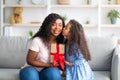 Mother& x27;s Day celebration. Lovely little black girl giving mom wrapped holiday gift and kissing her on cheek at home Royalty Free Stock Photo