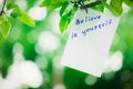 Motivating phrase Believe in yourself. On a green background on a branch is a white paper with a motivating phrase.