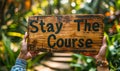 Motivational Stay The Course sign held up with two hands, promoting persistence and dedication, set against a lush green