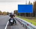 Motorcyclist standing on the highway in front of the blank information board with blue background, autumn season