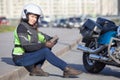 Motorcyclist woman in white crash helmet looking at camera, holding smartphone while sitting on roadside near chopper motorcycle Royalty Free Stock Photo