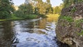 A motorized plastic boat floats on the river and creates a wave. One bank is reinforced with stone and the other with logs, on whi