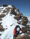 Mountain climber on a narrow rock and snow ridge on his way down from a high alpine summit with a metal summit cross in the backgr