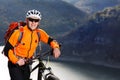 Mountain cyclist in the orange jacket stands under the beautiful river against mountains.