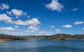 Blue calm deep water lake surrounded by hills with trees under a blue sky with small soft white clouds.