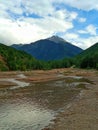 A mountain with a snow-capped peak is visible from a small plateau through which a small river flows. Cloudy weather. Royalty Free Stock Photo