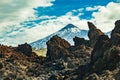 Mountain Teide with white snow spots, partly covered by the clouds. Bright blue sky. Huge lava rocks in the foreground. Teide