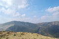 A grassy, rock strewn mountain top with other mountains beyond