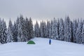 Mountain winter landscape. Tourist stands on the path next to the green tent. Wild forest. Touristic camping rest place.