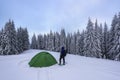 Mountain winter landscape. Tourist stands on the path next to the green tent. Wild forest. Touristic camping rest place.