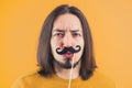 Movember concept - young frowned man holds fake paper moustache on a stick yellow background copy space isolated studio Royalty Free Stock Photo