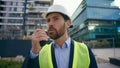 Moving shot Caucasian builder in hard hat helmet and safety vest talking in remote radio outdoors urban building