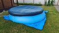 On a mown grass lawn near a wooden house there is a blue swimming pool, covered with a cover on top to avoid debris entering the w