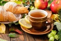 Mug of tea with lemon, croissant, autumn fall leaves, fruits on wooden background. Cozy home breakfast, autumn hygge style,
