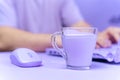 Mug with unusual lilac beverage and a computer mouse are on table, and person who works using a laptop on blurred Royalty Free Stock Photo