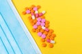 Multi-colored pills and a blue disposable mask on a bright yellow background, top view. Medication for the disease. Drugs from cor