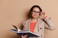 Charming middle-aged woman teacher holding a book with fairy tails, smiling at camera through spectacles, beige backdrop