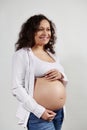 Multi ethnic curly gravid woman holding hands on her belly, smiling cutely looking away, isolated over white background