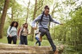 Multi ethnic group of five young adult friends running in a forest and jumping over fallen tree during a hike