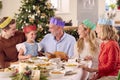 Multi-Generation Family Celebrating Christmas At Home Wearing Paper Hats Before Eating Meal Together