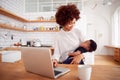 Multi-Tasking Mother Holds Sleeping Baby Son And Works On Laptop Computer In Kitchen Royalty Free Stock Photo