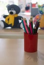 Multicolored pencils in a red cup on a wooden table. In the background - children`s toys. Children`s corner - a place for drawin