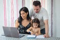 Family gathered at the dining room table, mother at the laptop, father standing hugging them and baby girl painting in a notebook Royalty Free Stock Photo