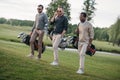 Multiethnic friends holding bags with golf clubs and walking on golf course Royalty Free Stock Photo