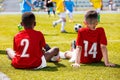 Two Young Multiracial Boys Wearing Red Soccer Jerseys Sitting on Grass at Soccer Field and Supporting Teammates